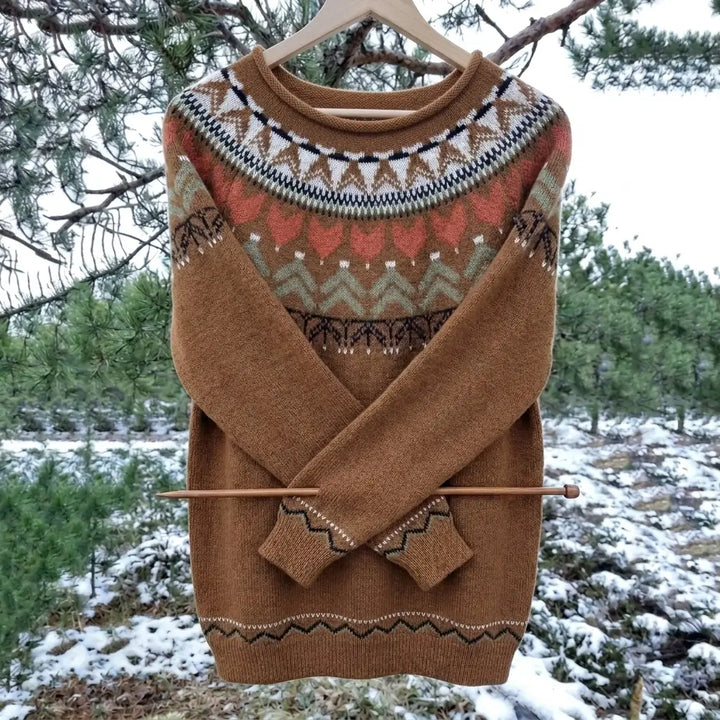 A brown knit sweater with a colorful Fair Isle yoke pattern, hanging on a wooden hanger outdoors on tree branches with snow in the background.